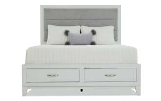 Twin bed with white wooden frame and gray upholstered headboard, featuring a white quilted bedspread, decorative pillows, and a built-in storage drawer.