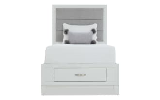 Twin bed with white wooden frame and gray upholstered headboard, featuring a white quilted bedspread, decorative pillows, and a built-in storage drawer.