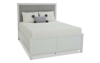 Queen-size bed with a light gray upholstered headboard, white panel footboard, and white bedding with decorative pillows, modern design.
