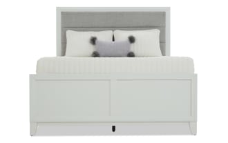 Queen-size bed with a light gray upholstered headboard, white panel footboard, and white bedding with decorative pillows, modern design.