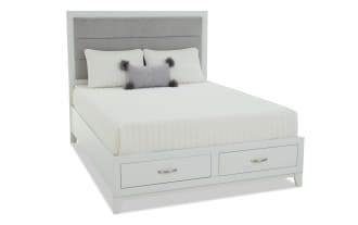 Modern white wooden bed frame with built-in storage drawers, upholstered gray fabric headboard, and cream bedding with decorative pillows.