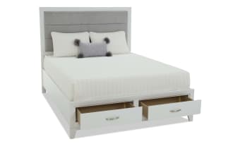 White bedroom furniture set including a bed with storage drawers, a dresser with mirror, a nightstand with lamp, and a tall chest, all in modern white wood finish.
