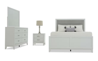 White bedroom furniture set including a bed with paneled headboard and footboard, a six-drawer dresser with mirror, and a two-drawer nightstand with a lamp, all in a modern, clean-lined design.