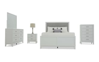 White bedroom furniture set including a bed with gray upholstered headboard, white dresser with mirror, nightstand with lamp, and tall chest, all with silver handles.