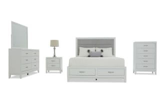 White bedroom furniture set including a bed with storage drawers, gray upholstered headboard, nightstand with lamp, dresser with mirror, and tall chest, all with silver handles.