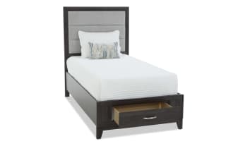 Dark wood bedroom set with a dresser and mirror, two-drawer nightstand with silver lamp, and a twin bed featuring a gray upholstered headboard and storage drawer.