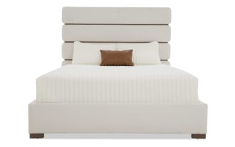 Modern bedroom furniture set with a cream upholstered bed featuring a padded headboard, wooden dresser with mirror, matching nightstand with lamp, and tall chest of drawers in walnut finish.