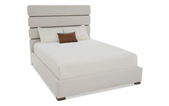 Modern upholstered bed with a beige fabric frame and headboard featuring horizontal panel design, white quilted bedding, and a brown accent pillow.
