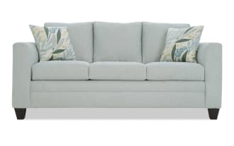 Light blue fabric sofa with three seat cushions, three back cushions, dark wooden legs, and two decorative floral-patterned throw pillows.