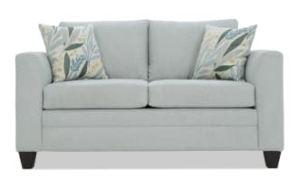 Light blue fabric loveseat sofa with two floral patterned throw pillows, featuring cushioned armrests and dark wooden legs, modern style.