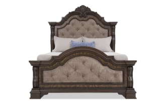 King-size bed with dark wood frame and ornate carved details, featuring tufted beige upholstered headboard and footboard, styled with neutral bedding and a blue accent pillow.