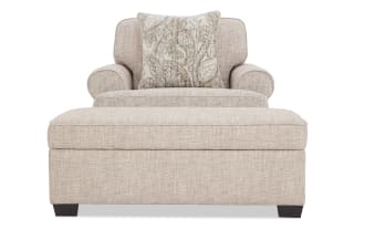 Beige upholstered armchair with matching ottoman, featuring a textured fabric, rounded arms, cushioned back, and decorative paisley-patterned throw pillow.