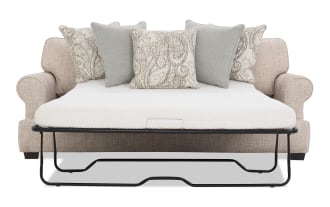 Beige fabric sofa bed with rolled arms, featuring a white mattress and five decorative pillows in beige paisley and solid gray patterns, supported by a black metal frame.