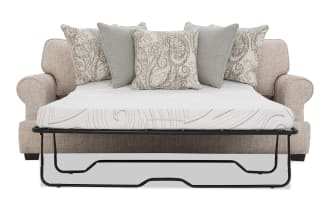 Beige fabric sofa bed with rolled arms, featuring a black metal frame fold-out mattress with a white quilted cover and five decorative throw pillows in beige and gray patterns.
