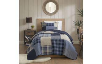 Twin bed with blue and beige patchwork quilt and matching pillow, wooden headboard, light wood panel walls, and cozy room decor.