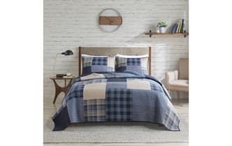 Modern bedroom with a wooden bed frame and beige upholstered headboard, featuring a blue and beige plaid quilt set, matching pillow shams, a mid-century style nightstand with a black lamp, a round wall clock, floating shelf with books and plant, light gray armchair, and light wood flooring with a gray area rug.