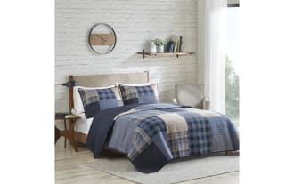 Modern bedroom with a wooden bed frame and beige upholstered headboard, featuring a blue and beige plaid quilt set, matching pillow shams, a mid-century style nightstand with a black lamp, a round wall clock, floating shelf with books and plant, light gray armchair, and light wood flooring with a gray area rug.