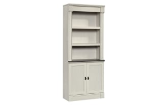 Tall gray wooden bookshelf with two lower cabinet doors, open shelving with decorative items including books, vases, and framed photo, classic design.