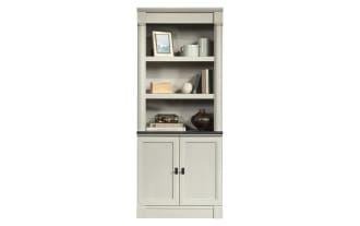Tall gray wooden bookshelf with two lower cabinet doors, open shelving with decorative items including books, vases, and framed photo, classic design.