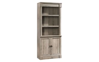 Tall wooden bookshelf and cabinet unit in light weathered gray finish with two open shelves, decorative items, and two-door storage below.