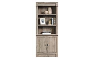 Tall wooden bookshelf and cabinet unit in light weathered gray finish with two open shelves, decorative items, and two-door storage below.