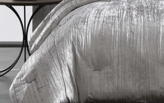 Luxurious silver crushed velvet comforter set with matching decorative pillows on a gray upholstered bed frame, styled in a modern bedroom.