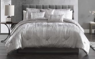 Luxurious silver crushed velvet comforter set with matching decorative pillows on a gray upholstered bed frame, styled in a modern bedroom.
