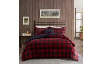 Bed with black metal frame, red and black buffalo plaid comforter set including matching pillowcases and a navy blue throw pillow with bear embroidery, against a wood-paneled wall with a framed landscape artwork.