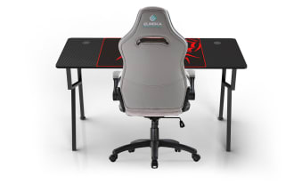 Modern gaming setup with a black carbon fiber textured desk featuring dual monitors, mechanical keyboard, mouse, and speakers. Includes a gray and white ergonomic EUREKA ergonomic gaming chair with padded armrests and adjustable height on caster wheels. Desk has a large black and red scorpion-themed mouse pad.