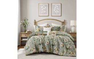 Bed with green floral-patterned comforter and matching pillows, wooden headboard, beige rug, and natural light from large window in cozy bedroom.