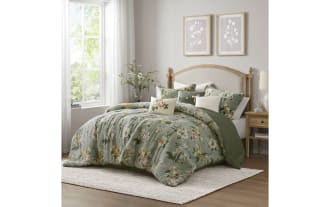 Bed with green floral-patterned comforter and matching pillows, wooden headboard, beige rug, and natural light from large window in cozy bedroom.
