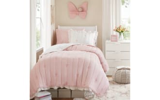 White and pink twin bed set with ruffled edges and embroidered bows, featuring a comforter, pillow shams, and floral accent pillow in a cozy bedroom setting.