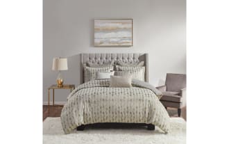 Queen-size bed with tufted gray upholstered headboard, patterned beige and gray comforter set, matching pillows, gold glass table lamp on brass side table, gray armchair, and abstract wall art.
