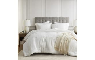 King-size bed with a tufted gray fabric headboard, white textured bedding, matching pillows, cream throw blanket, wooden nightstands, and ceramic lamps.