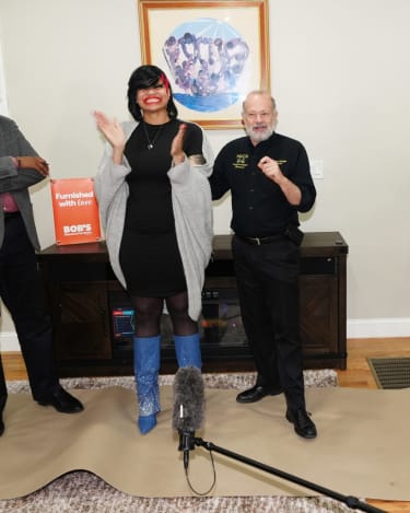 Two people standing indoors near a dark wood fireplace console; one wears a black dress, gray shawl, and blue embellished boots, the other wears black pants and a black shirt. A sign on the console reads "Furnished with love BOB'S Discount Furniture." A microphone boom is visible in the foreground.
