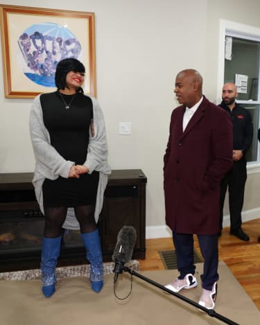 Two people standing indoors near a fireplace and window; one wearing a black dress, gray cardigan, and blue embellished boots, the other in a maroon coat, navy pants, and white sneakers. A microphone on a boom pole is positioned in front of them.