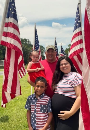 A family of four stands outdoors on grass surrounded by American flags, with the father holding a young girl in a red dress, a boy in a star-spangled shirt, and a pregnant woman in a striped top and black skirt, symbolizing patriotism and family unity.