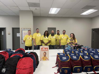 Group of seven volunteers wearing yellow "Bob's Volunteer" t-shirts standing behind tables filled with red, black, and navy backpacks and maroon and navy Drug Mart branded bags, with a sign reading "Bob's Outreach."