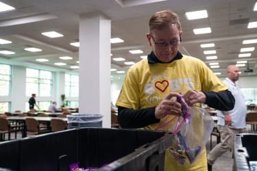 Volunteer wearing a yellow "Bob's Volunteers" shirt sorting candy into a clear plastic bag in a well-lit community center with tables and chairs.