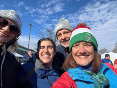 Five people wearing winter clothing and hats, smiling outdoors under a partly cloudy sky, with tents and a building in the background.