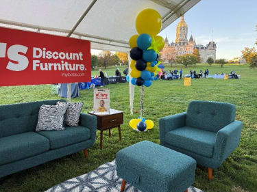 Outdoor event seating area with teal fabric sofa, armchair, ottoman, zebra-patterned pillows, and a side table under a white canopy. Red banner reads "Discount Furniture. mybobs.com." Yellow, blue, and black balloon arrangement nearby. Historic building and green lawn in background.