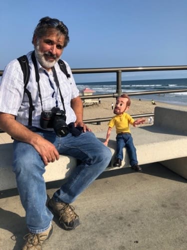 Man wearing a striped shirt and jeans with a Nikon camera around his neck sitting on a concrete bench by the beach next to a small doll in a yellow shirt and jeans.