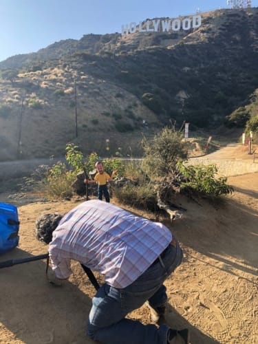 Man kneeling on a dirt path taking a photo of a manikin figure with a beard and yellow shirt near the Hollywood sign on a sunny day.