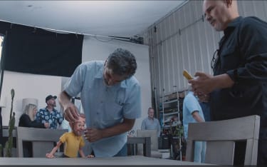 A man in a light blue checkered shirt adjusts a small articulated doll wearing a yellow polo shirt on a table in a workshop setting with other people and equipment in the background.