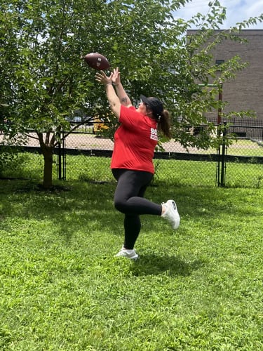 Woman in a red shirt and black leggings catching a football outdoors on green grass near a tree and chain-link fence.