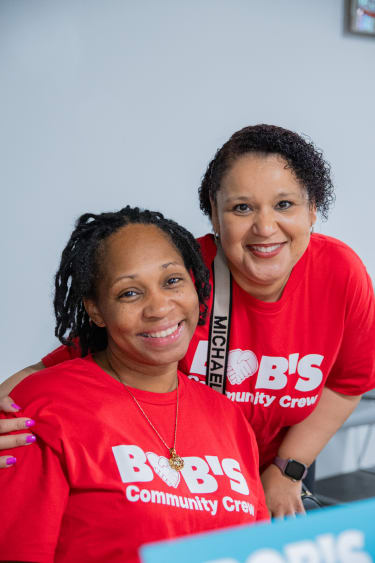 Two smiling women wearing red "BOB'S Community Crew" t-shirts, showing team spirit and community involvement.