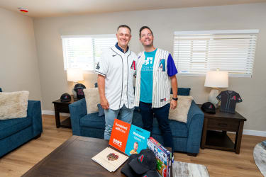 Two men wearing Arizona Diamondbacks baseball jerseys stand smiling in a furnished living room with blue sofas, wooden side tables, and table lamps. On the coffee table are promotional materials from Bob's Discount Furniture, including a card that reads "Furnished with love" and "Welcome home," alongside a baseball-themed board game and an Arizona Diamondbacks cap.