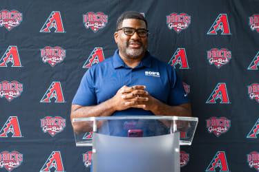 Man wearing glasses and a blue polo shirt with "BOB'S Discount Furniture" logo, standing behind a clear podium with a backdrop featuring Arizona Diamondbacks logos and "D-Backs Give Back" text.