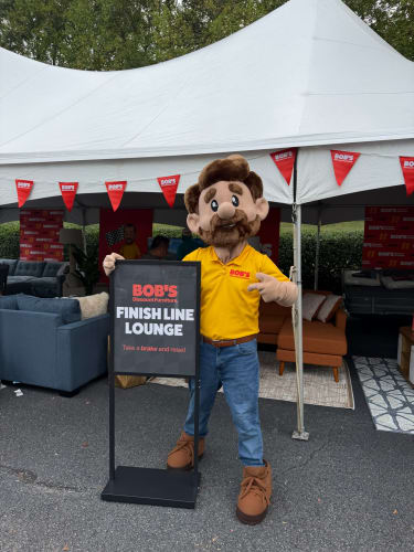 Mascot wearing yellow Bob's Discount Furniture shirt stands next to sign reading "BOB'S Discount Furniture. FINISH LINE LOUNGE. Take a brake and relax!" under event tent with sofas and chairs.