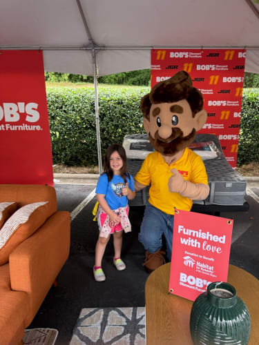 A young girl stands next to a person in a Bob's Discount Furniture mascot costume under a tent. Behind them is a mattress and a red sign reading: "Furnished with love. Donation to benefit Habitat for Humanity of The Charlotte Region. BOB'S Discount Furniture." The setting appears to be a promotional event for Bob's Discount Furniture supporting Habitat for Humanity.